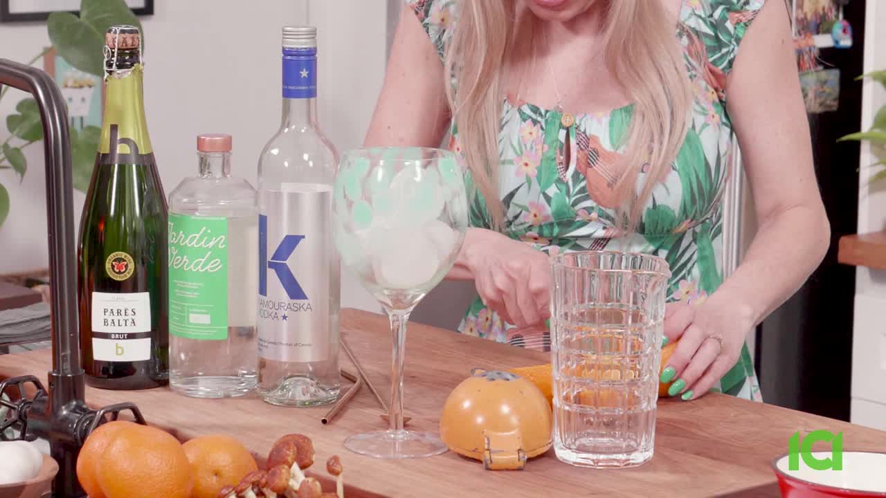A woman in a floral top slices an orange next to a tall, empty glass. Bottles of liquor and champagne sit behind her on the wooden counter.
