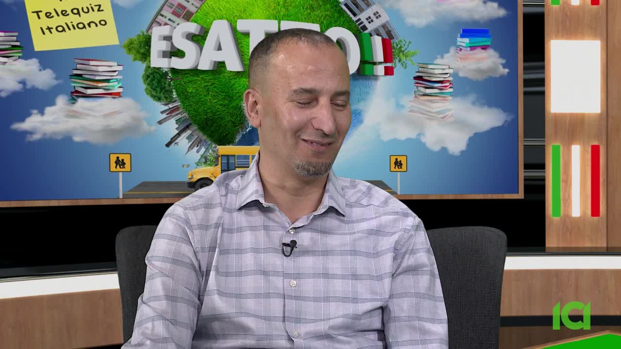 The man in the checkered shirt smiles slightly, seated in front of a backdrop with the "ESATTO!" logo and a cartoonish school bus. The CFHD-DT studio in Canada is clearly the setting for this Italian-language quiz show.
