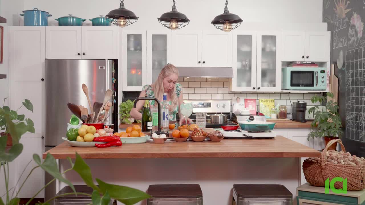 A woman in a floral top is busy at the kitchen island, pouring something from a bottle. On the stove, a pan bubbles gently, while a variety of ingredients surround her.

