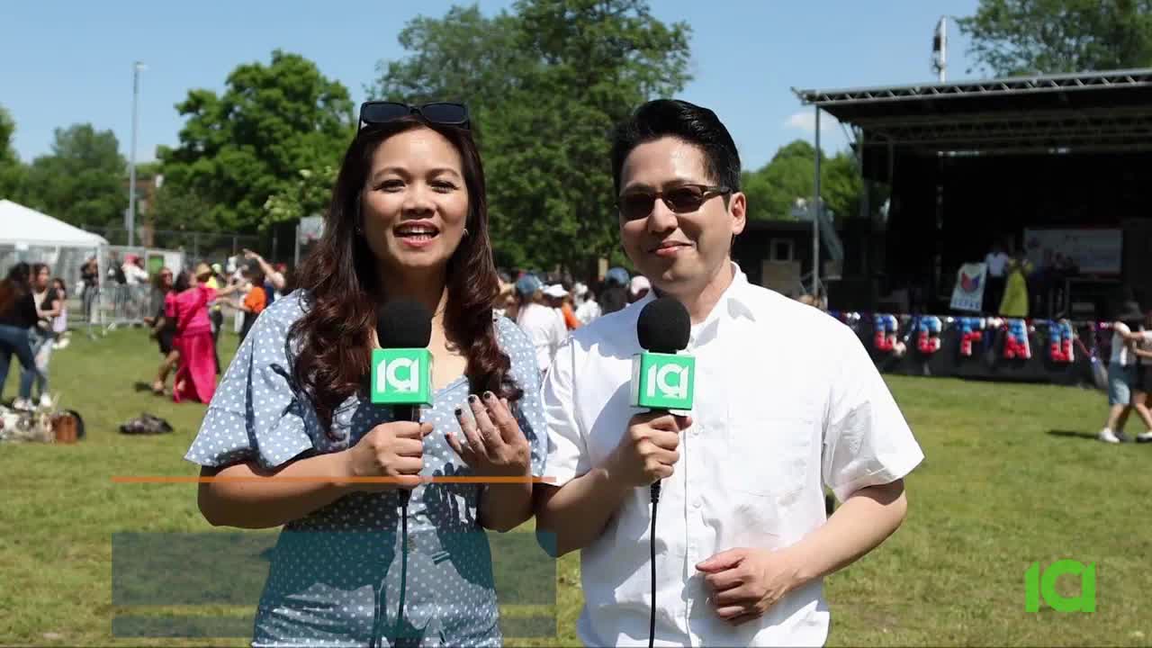 Two CFHD-DT reporters stand in front of a crowd, holding microphones with the station's logo. Behind them, people are gathered on the grass, and a stage is set up for a performance.

