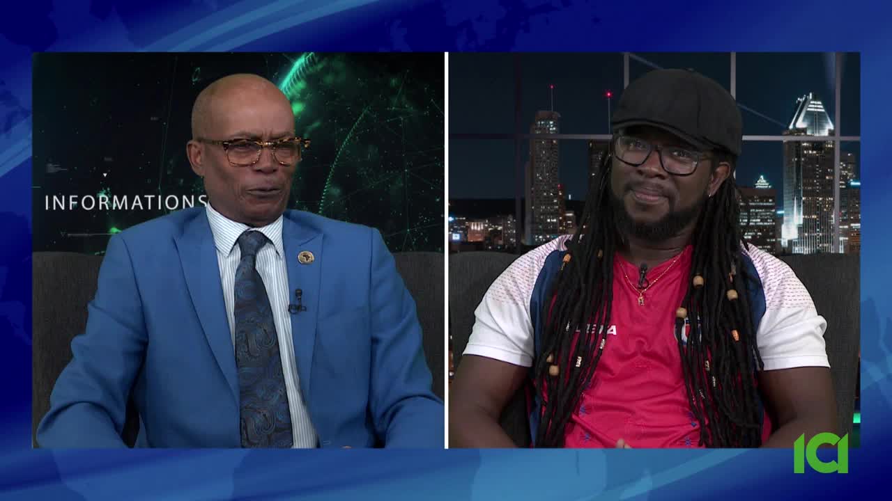 A man in a blue suit and glasses is speaking, while another man with dreadlocks and a cap listens. The scene appears to be a CFHD-DT studio in Canada, with a city skyline visible in the background.
