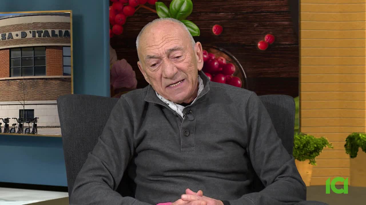 A man in a gray sweater is speaking, his mouth moving as he gestures with his hands. Behind him, a backdrop on CFHD-DT features a framed image and some decorative elements.
