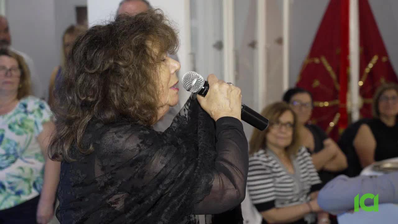 A woman with curly hair sings into a microphone, her black shawl draped over her arm. Behind her, a group of people watches, some smiling, as the CFHD-DT broadcast unfolds.
