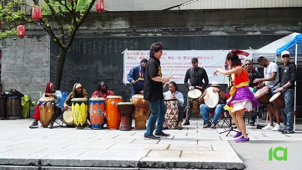 A group of drummers sits behind a woman in a colorful dress, who dances in front of them. The man in the black shirt seems to be directing the performance.
