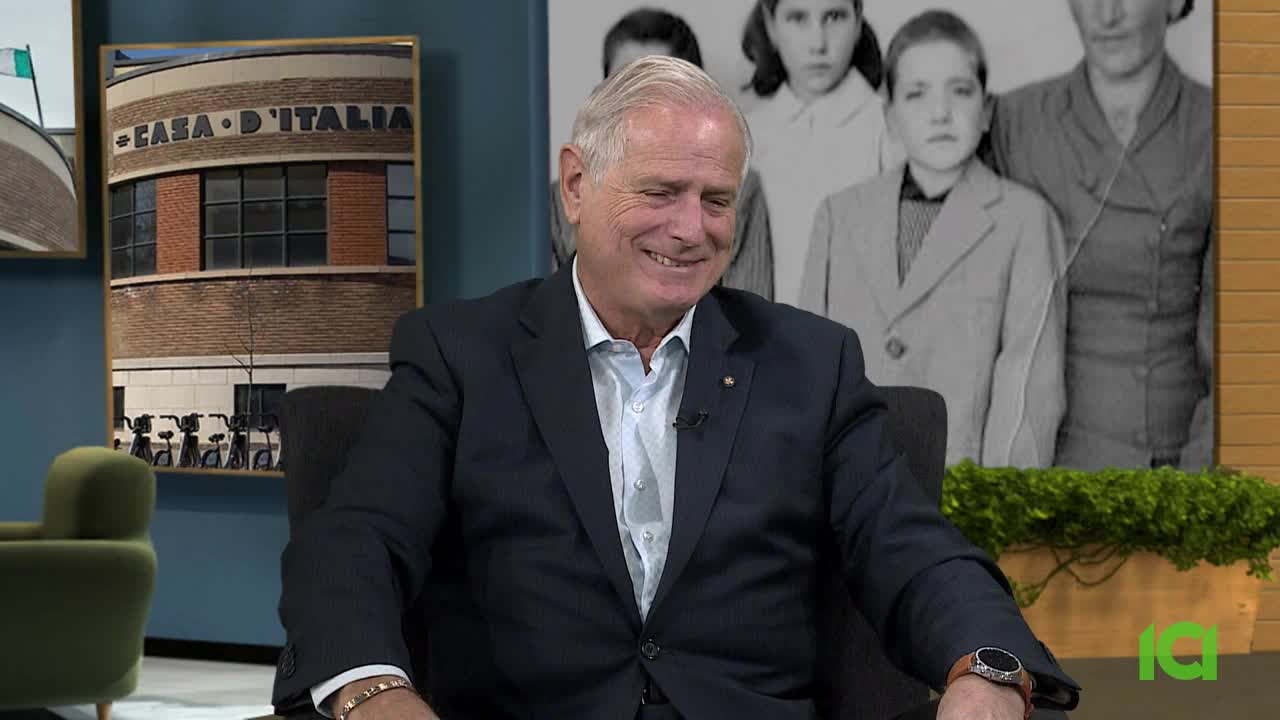 A man in a suit sits in a chair, smiling at someone off-camera. Behind him, a framed picture of a building labeled "Casa D'Italia" hangs on the wall, and a black and white photo of a family.
