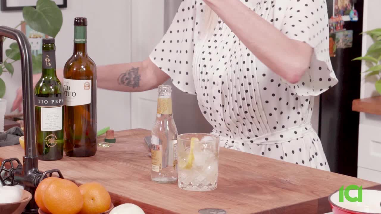 A woman in a polka-dot dress is pouring something from a bottle into a glass of ice and lemon. Two bottles of wine and a bottle of tonic water sit on the wooden countertop beside her.
