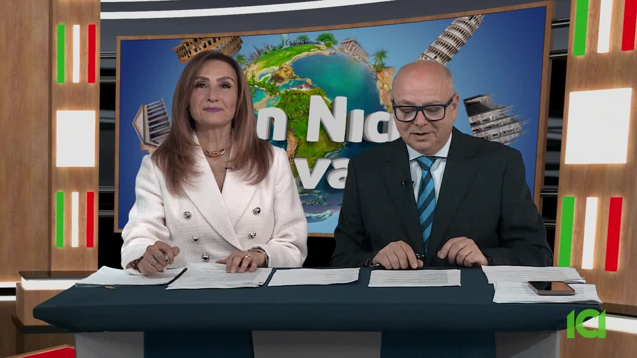 Two people sit behind a desk, reading from papers. Behind them, a screen displays a globe and the words "San Nicola Viva" in front of a backdrop with the colors of the Italian flag, likely a CFHD-DT broadcast from Canada.
