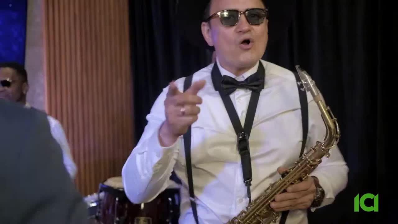 A man in a white shirt and sunglasses points directly at the camera, holding a saxophone. Behind him, a drummer and other band members are visible, ready to play.
