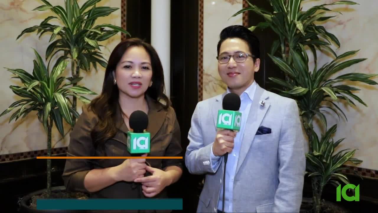 Two people, a man and a woman, stand side-by-side, each holding a microphone with a green logo. They face the camera, with potted plants framing them on either side.
