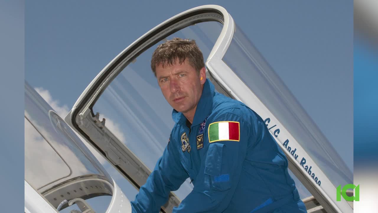 A man in a blue flight suit leans against the open cockpit of a plane, the Italian flag patch prominent on his shoulder. He looks directly at the camera, with a serious expression, under a clear, blue sky.
