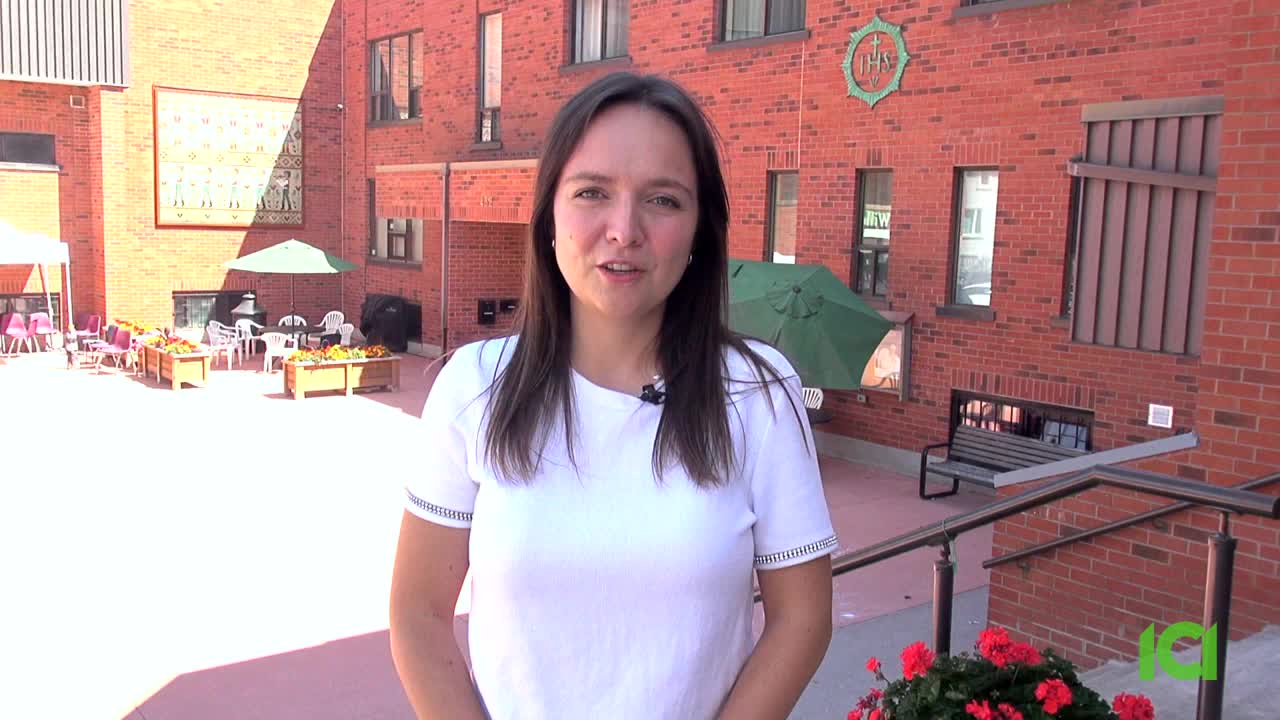 A woman in a white shirt speaks directly to the camera, a small microphone clipped to her collar. Behind her, a brick building and patio furniture sit in the bright sunshine.

