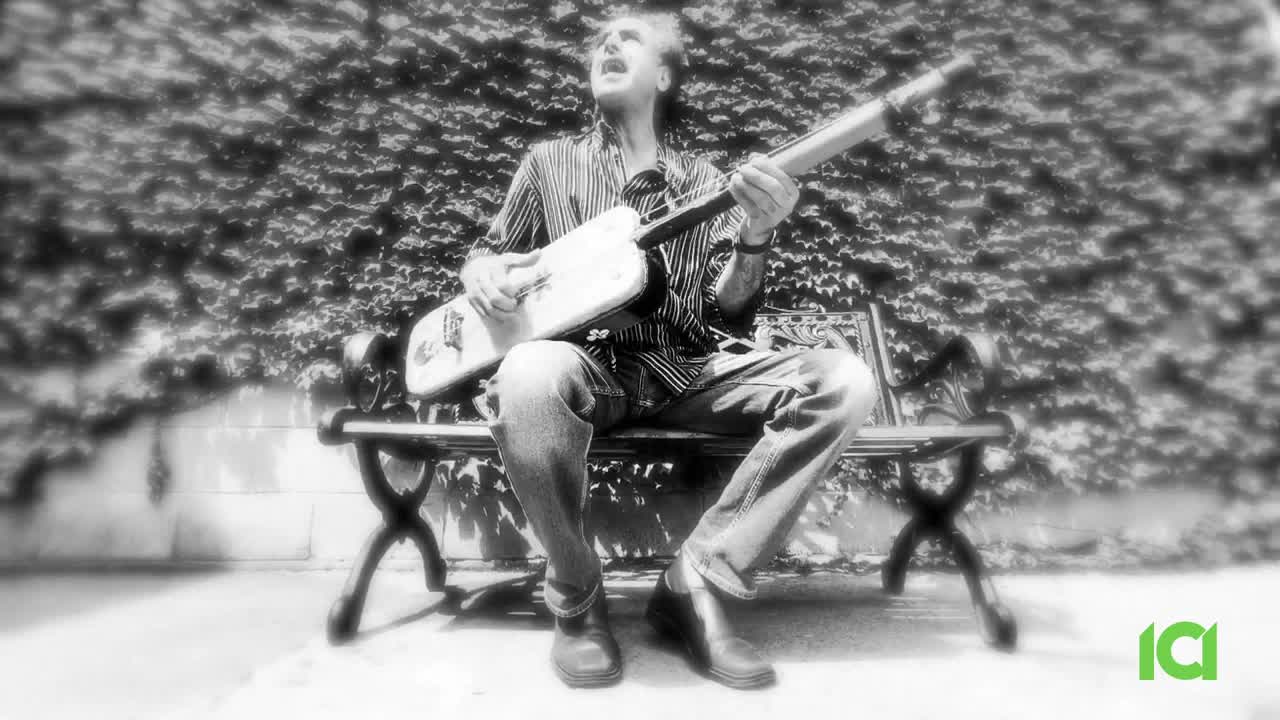 A man sits on a bench, playing a guitar and singing with his mouth wide open. The musician is outside, with a leafy wall behind him, as CFHD-DT broadcasts from Canada.
