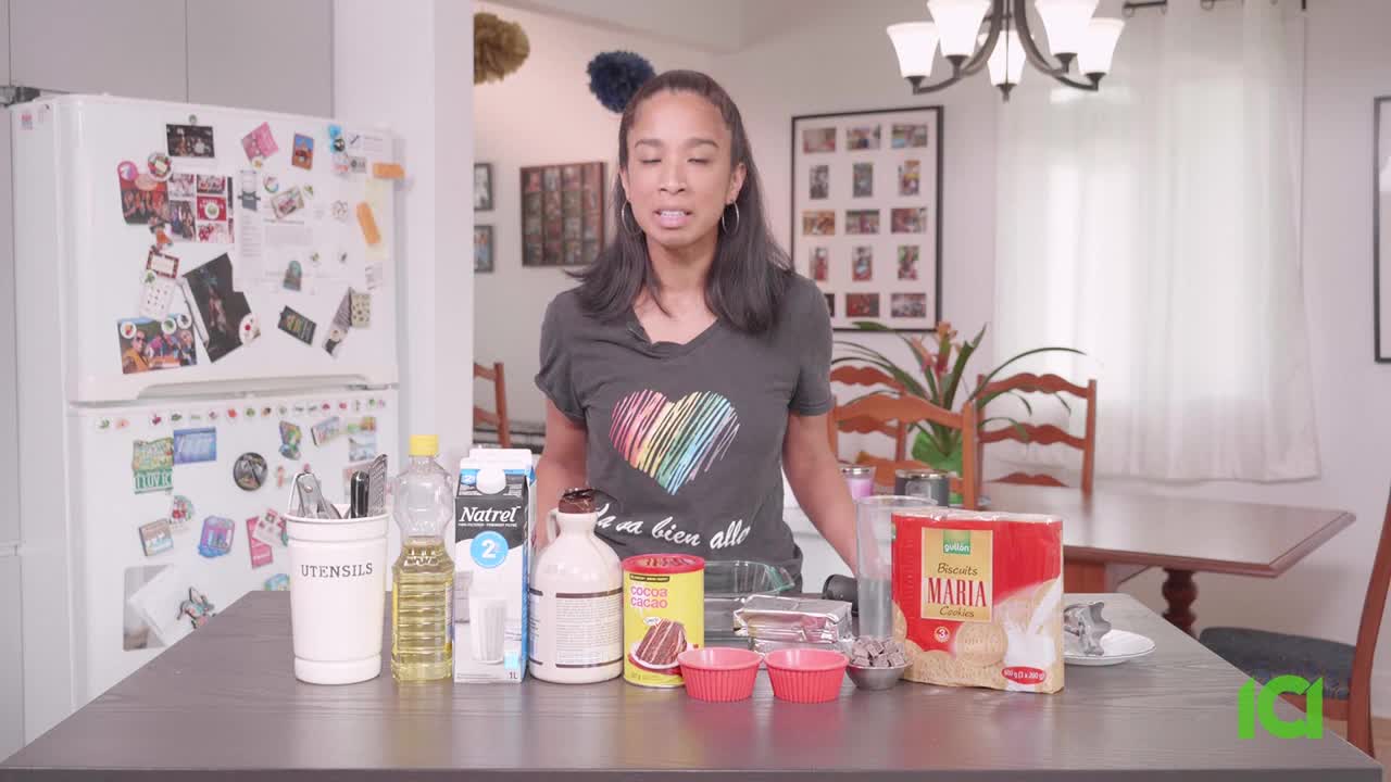 A woman in a heart-adorned shirt stands in front of a table laden with ingredients. She looks directly at the camera, likely about to begin a cooking demonstration for CFHD-DT in Canada.
