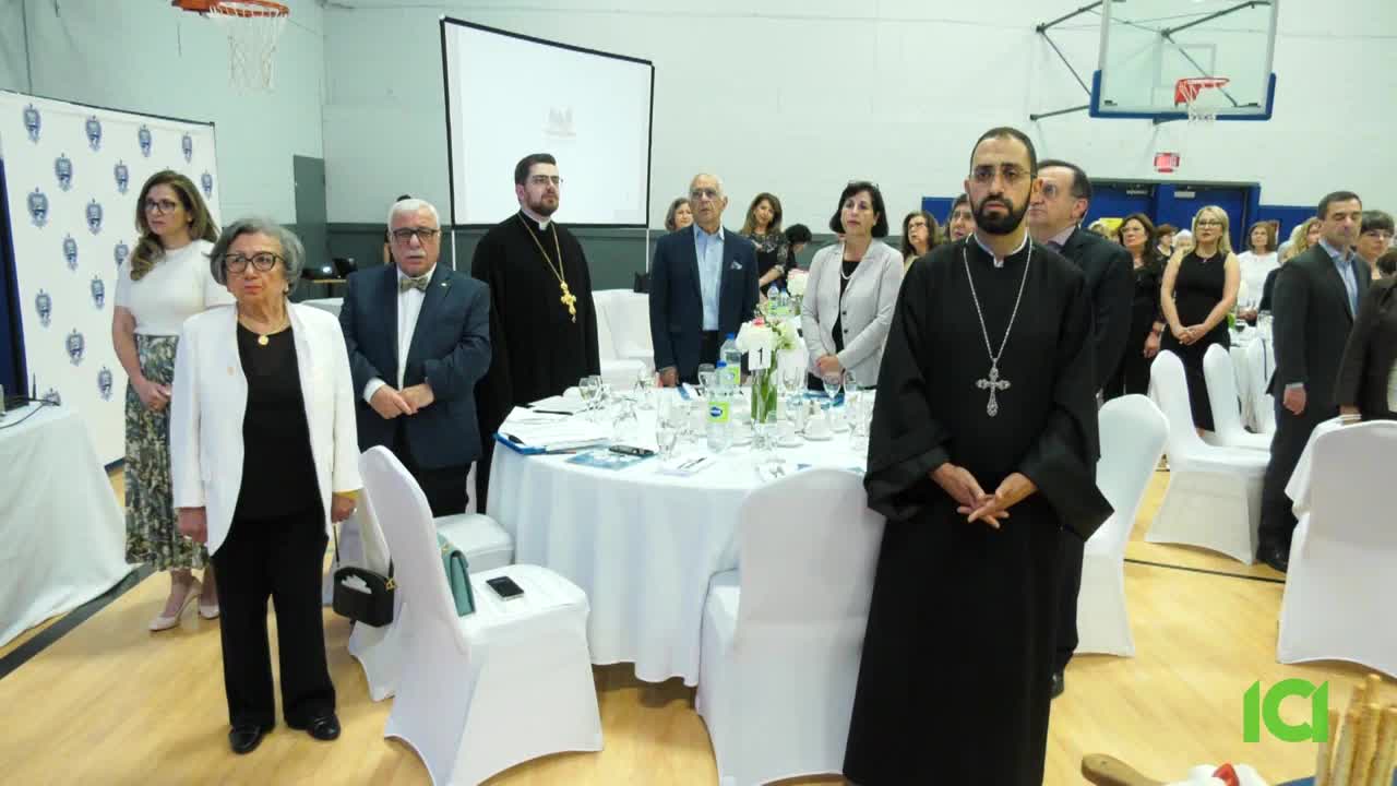 People stand at attention in a gymnasium, some in formal attire and others in religious garb. A table is set with a centerpiece, and a screen displays a logo.
