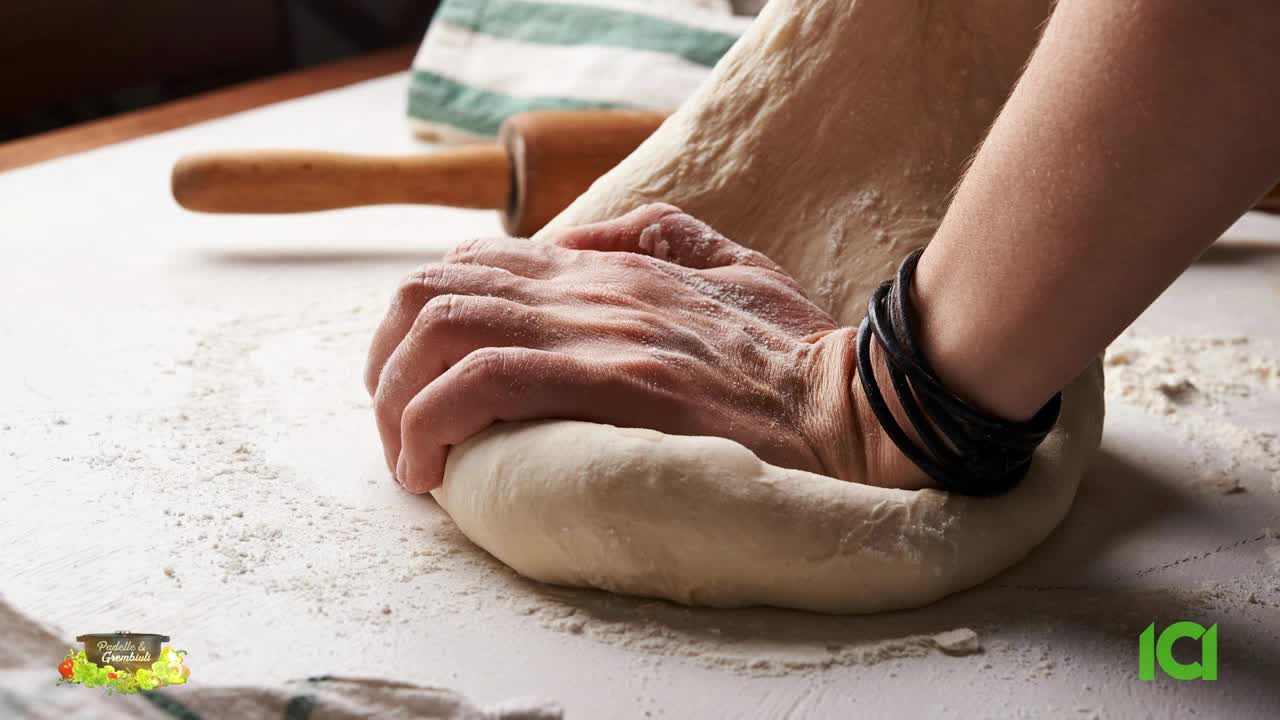 Hands are pressing into a mound of dough, kneading it on a floured surface. A wooden rolling pin rests nearby, and a towel is folded in the background.
