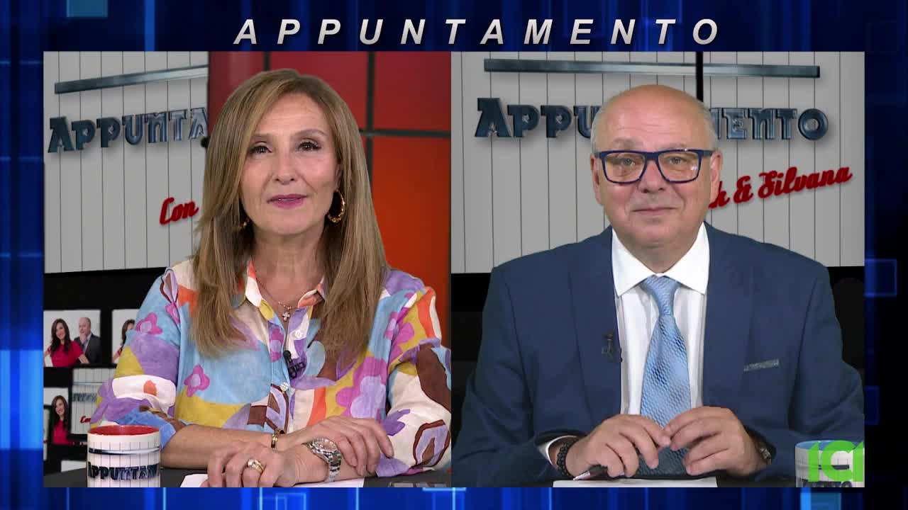 Two people sit at separate desks, facing the camera. The woman on the left wears a colorful patterned shirt, while the man on the right is in a blue suit and tie.
