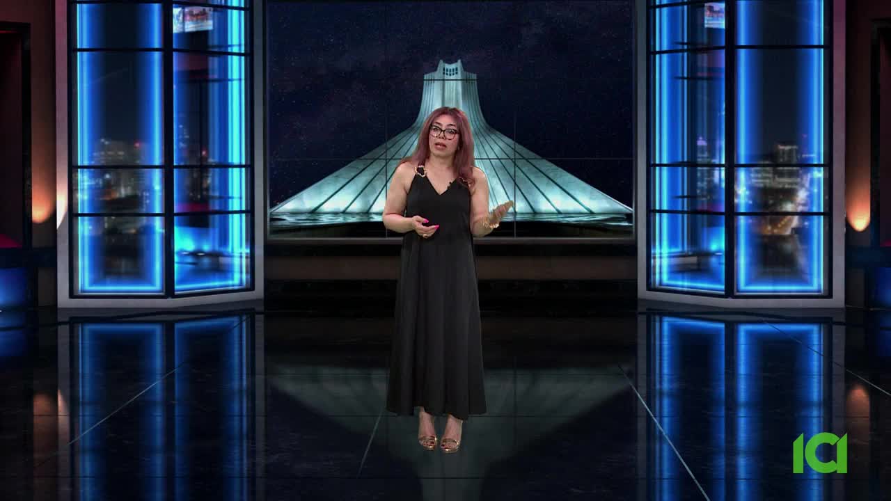 A woman in a black dress gestures with her hands, speaking in front of a backdrop featuring a stylized building. The set is lit with blue neon, reflecting on the glossy floor.
