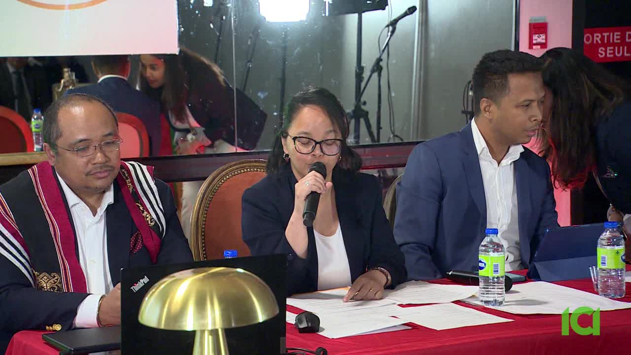 A woman in glasses speaks into a microphone, looking down at papers on the table in front of her. Two men sit beside her, one in a suit and the other wearing a patterned scarf, all facing the same direction.
