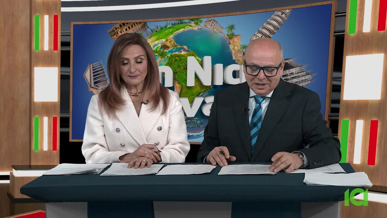 A man and woman sit behind a desk, reading from papers. The backdrop behind them features a globe and the words "San Nicola Viva."
