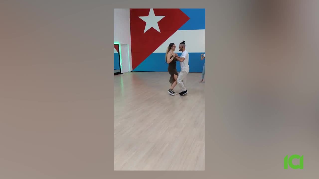 A couple dances in a studio, their bodies moving in sync. The man in white leads, guiding the woman in a dark dress across the wooden floor.
