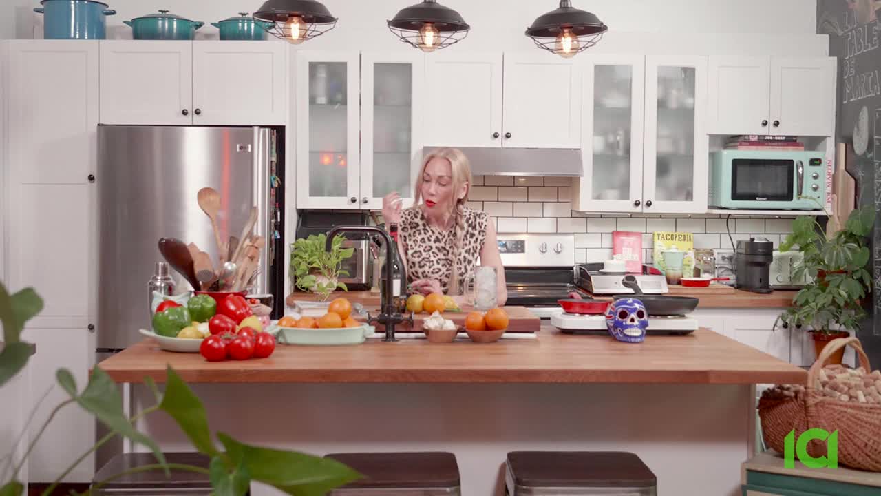 A woman in a leopard-print top gestures with her hand as she speaks, standing behind a kitchen island. The set is bright, with a colorful array of ingredients on the counter, and a skull sits next to a small pan.
