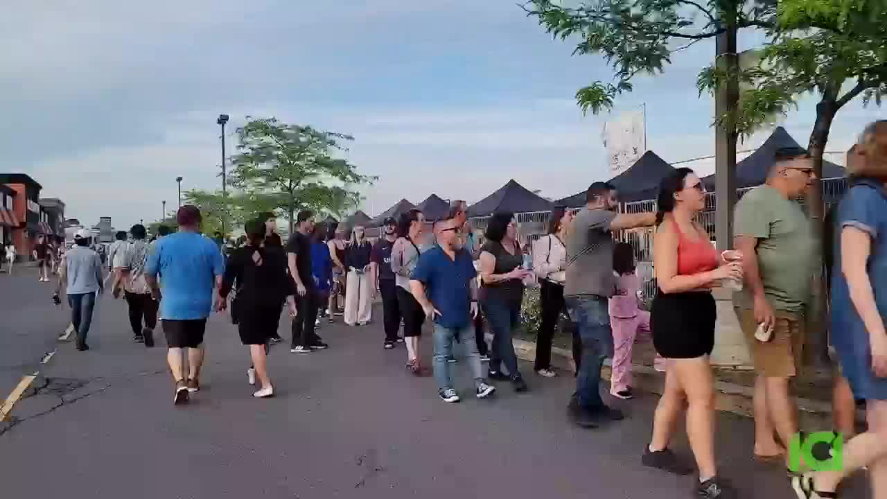 People are walking in both directions on a paved street, some in a long line. A few are holding drinks, and tents are set up in the distance.

