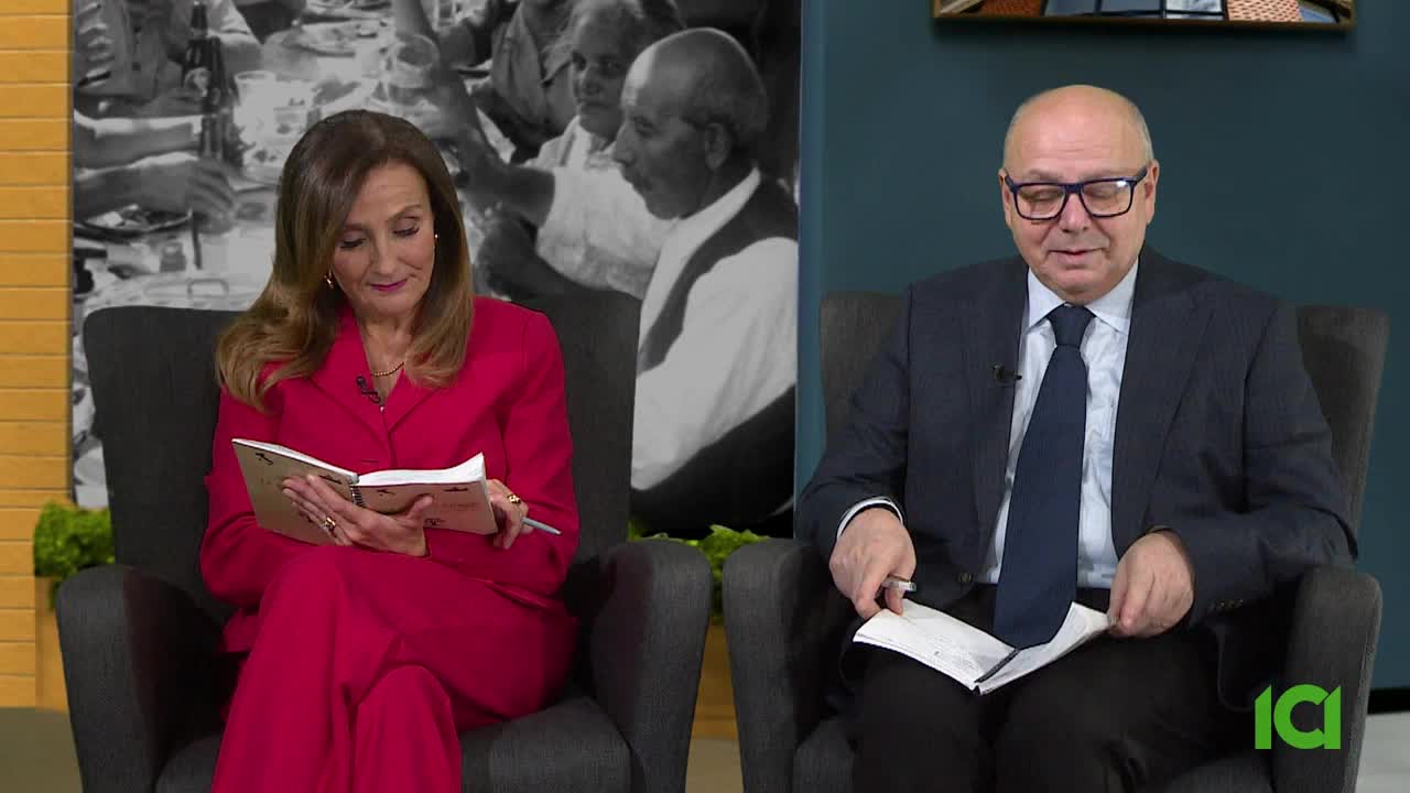 The woman in the red suit is looking down, reading from a notebook. Across from her, a man in a suit and tie holds a few papers, looking at them.
