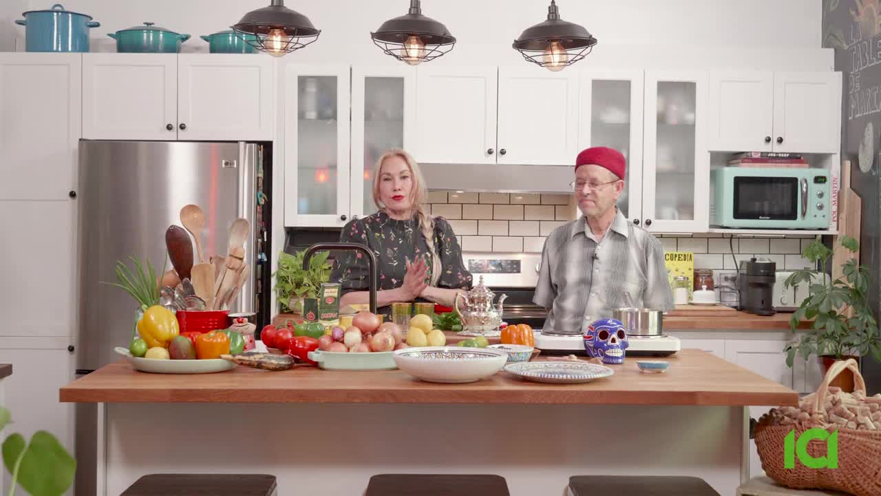 A woman and a man stand behind a wooden counter laden with fresh produce and cooking equipment. The woman gestures with her hands as the man, wearing a red fez, looks on.
