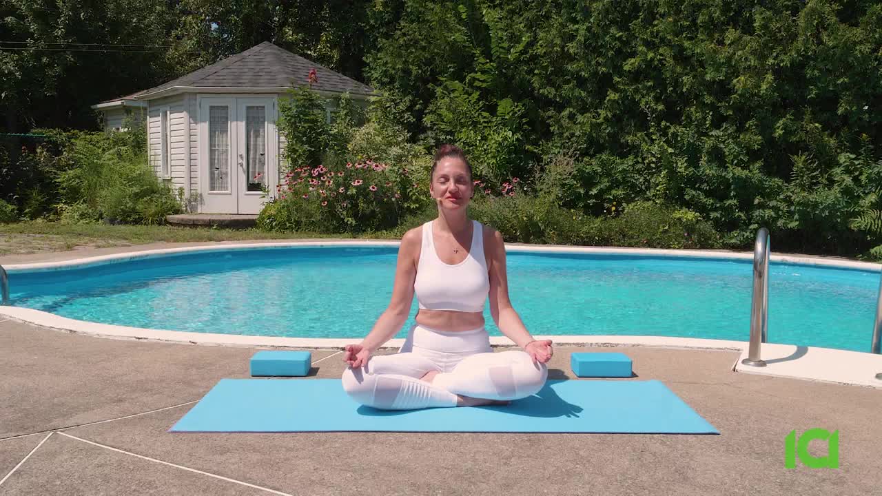 A woman in white workout clothes sits cross-legged on a blue mat beside a pool, eyes closed. Behind her, a small white building and lush greenery create a peaceful backdrop for what looks like a CFHD-DT broadcast from Canada.
