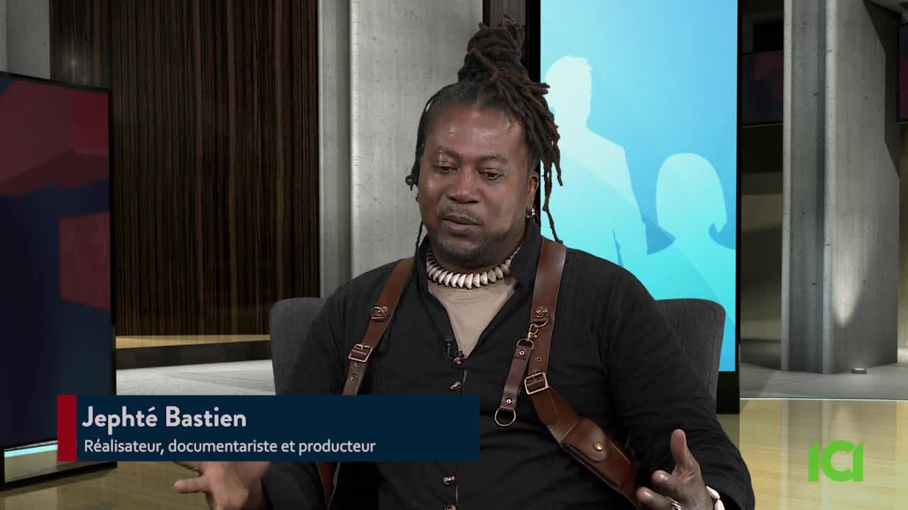 A man with dreadlocks and leather camera straps is seated, speaking on a television set. A lower third identifies him as Jephthé Bastien, a director and documentarian.
