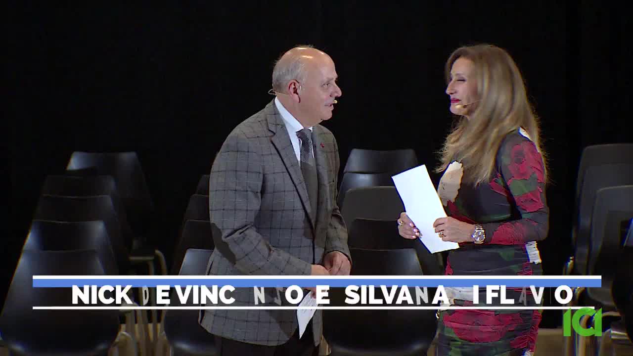 The man in the checkered jacket is speaking, gesturing slightly toward the woman holding papers. They're on a stage, with rows of empty chairs behind them, and the CFHD-DT logo is visible.
