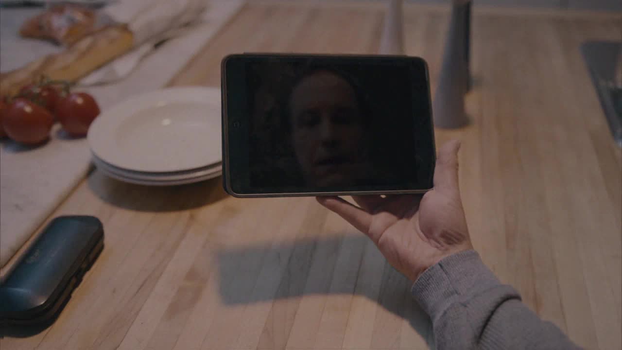 A person's face is reflected in the dark screen of a tablet, held out over a wooden countertop. Beside the tablet, a stack of plates and some tomatoes sit on the counter.
