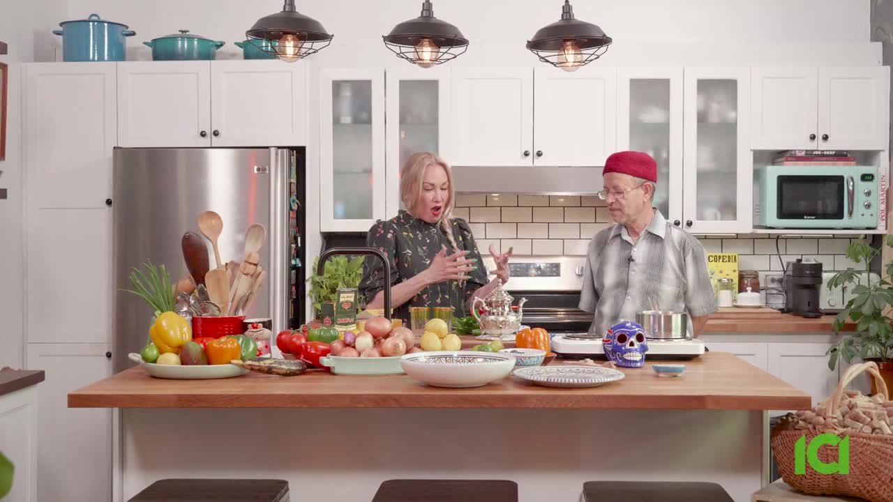 A woman gestures with a wooden spoon while speaking, the other man stands beside her, looking at a pot on a burner. A table laden with colorful vegetables and ingredients sits between them, in a bright kitchen.
