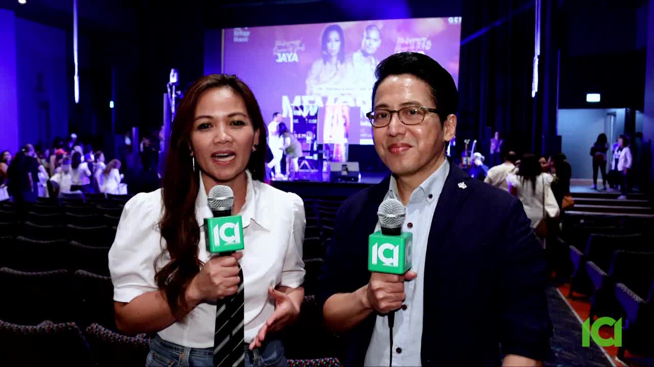 Two people with microphones stand in front of a large screen, speaking directly to the camera. Behind them, an audience mills about in a large auditorium, while a stage is set up in the distance.
