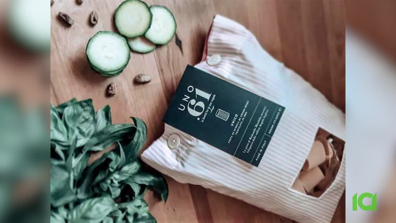 A package of pasta sits open, the contents visible through a cutout. Sliced zucchini, basil leaves, and a few nuts are scattered around it on a wooden surface.
