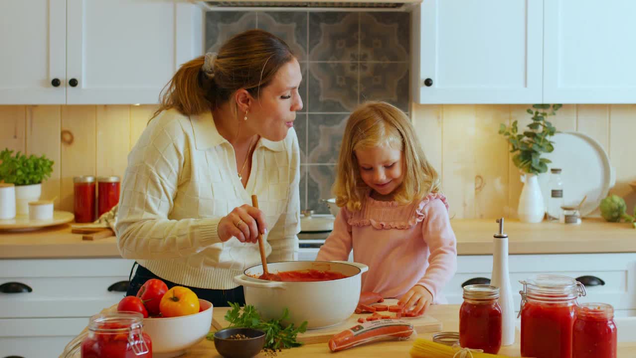 A woman, stirring a pot of red sauce, tastes it with a wooden spoon. Her daughter, focused on a cutting board, slices something small.

