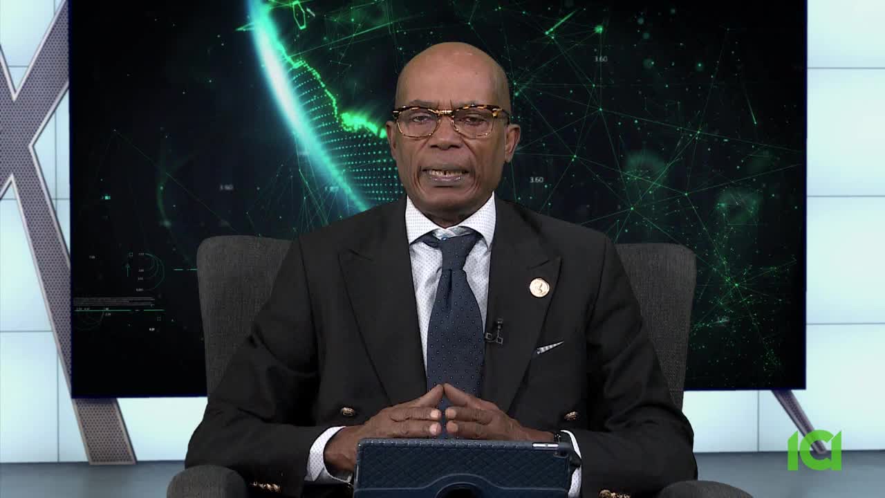 A man in a suit sits in a chair, speaking directly into the camera. Behind him, a digital graphic of a globe and interconnected lines fills a large screen.

