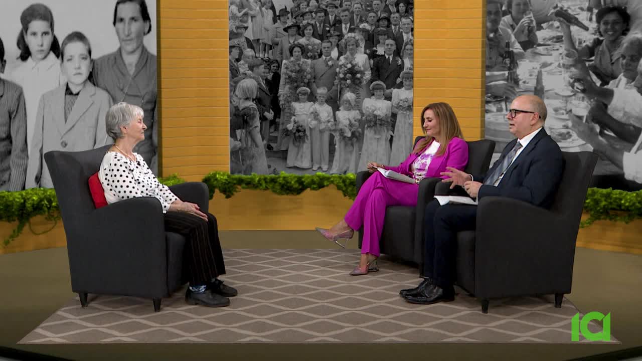 The woman in the polka-dot blouse sits in an armchair, facing the other two. The host in the pink suit gestures with her hands as the man in the suit speaks.
