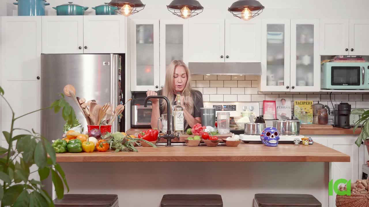 A woman in a kitchen is holding a bottle, looking at the camera, with a spread of ingredients laid out on the countertop. The scene is bright, with overhead lights and a well-stocked kitchen.
