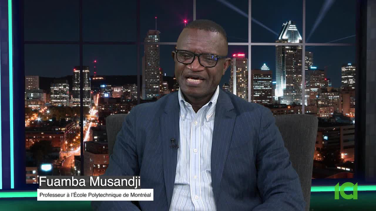 A man in a blue blazer and glasses is speaking, his face animated. Behind him, the night lights of a city in Canada shine through a large window.
