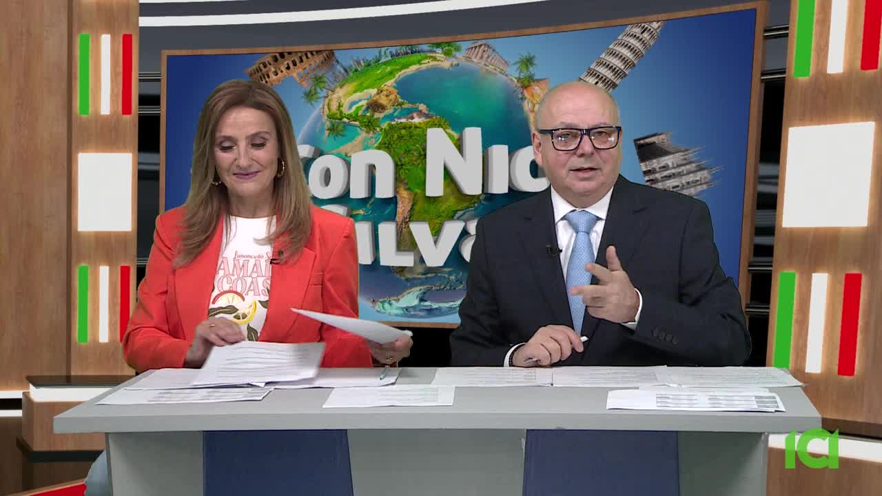 A woman in an orange blazer and a man in a suit sit behind a desk. The man gestures with his right hand, while the woman holds papers.

