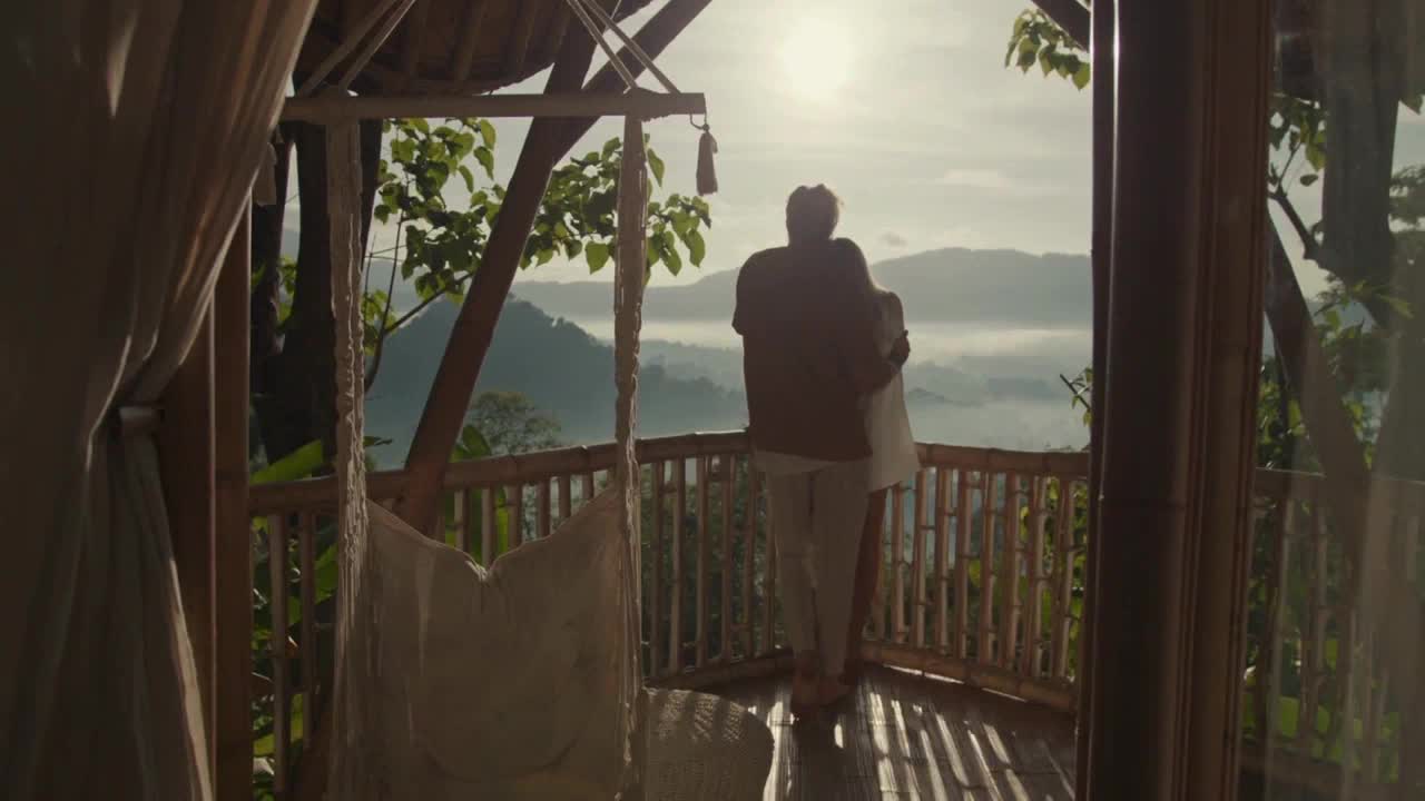 A couple stands on a wooden deck, arms wrapped around each other, gazing at a misty mountain vista. The sun shines brightly, illuminating the scene as if it were a CFHD-DT broadcast from Canada.
