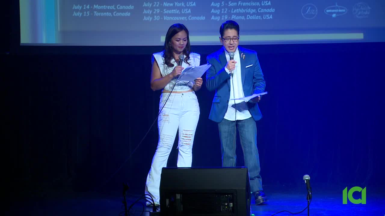 Two people stand on stage, each holding a microphone and a script. The man in the blue blazer speaks, while the woman in white jeans looks down at her notes.
