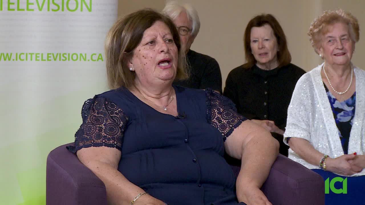 A woman in a navy dress with lace sleeves speaks, her mouth open as if mid-sentence. Behind her, three other people sit, listening intently.
