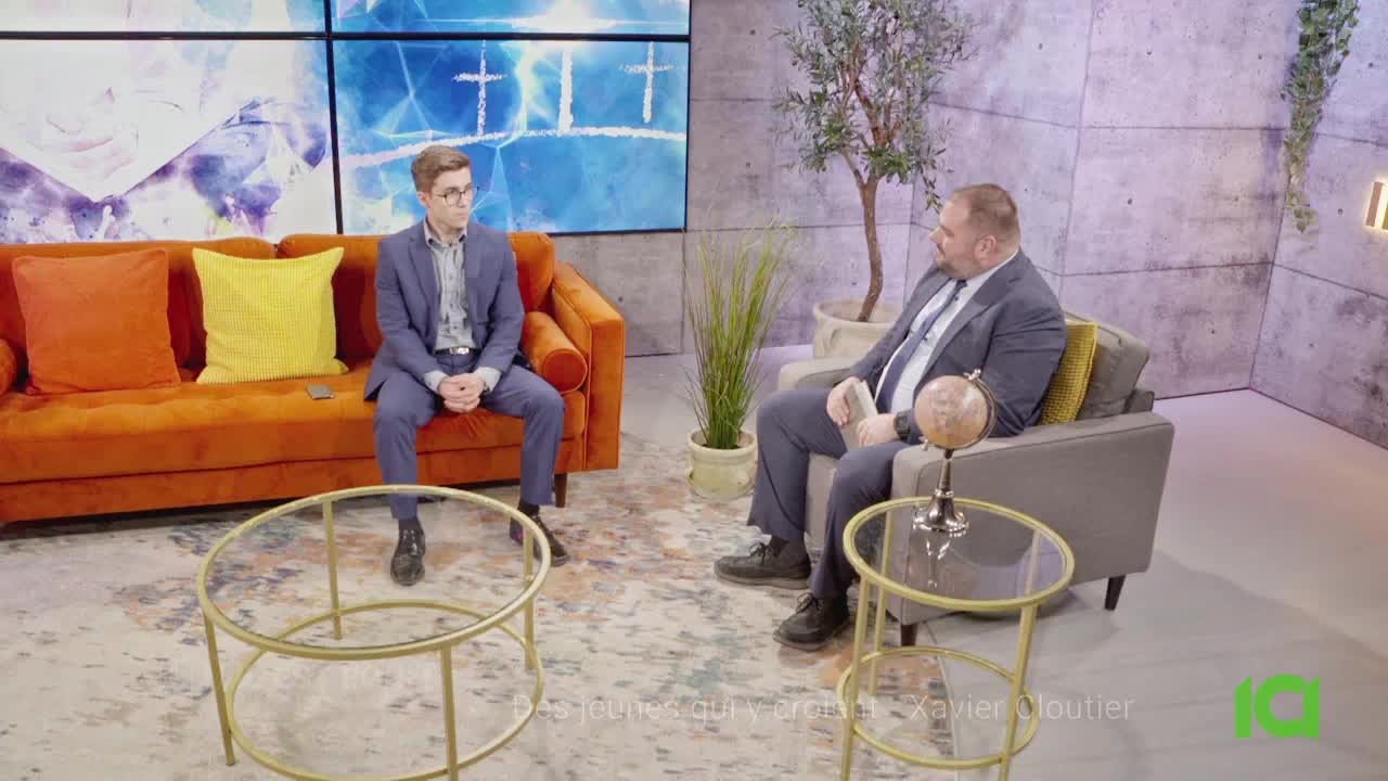 Two men in suits are seated, one on a burnt orange sofa, the other in an armchair, engaged in conversation. A glass-topped coffee table sits between them, and behind them, a large screen displays abstract art.
