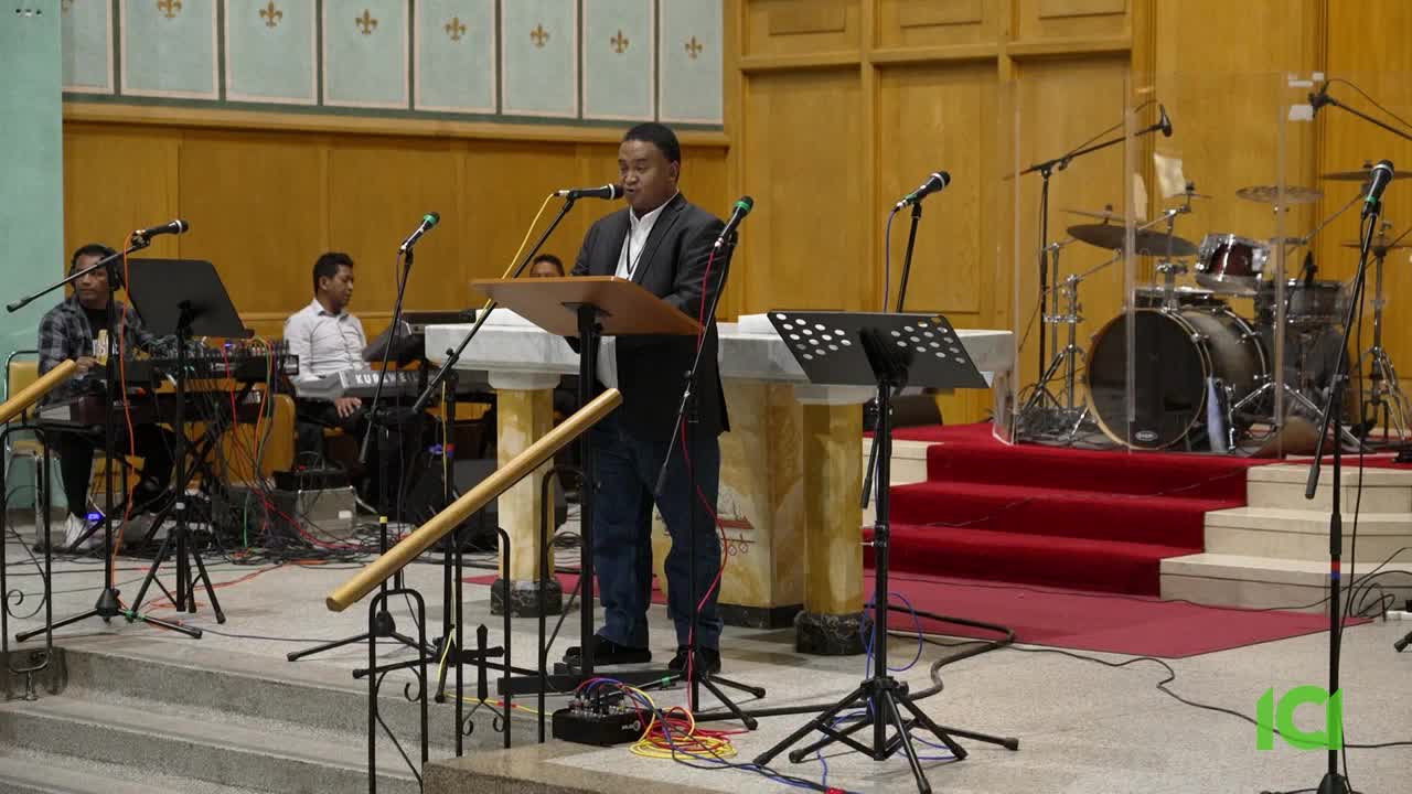 A man in a suit stands at a podium, speaking into a microphone. Behind him, a band is set up with keyboards, drums, and more microphones, ready to play.
