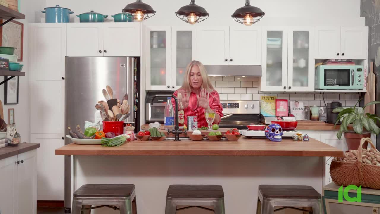 A woman in a red-striped shirt gestures with her hands, seemingly explaining something in a well-lit kitchen. Various ingredients and small bowls are arranged on a wooden countertop in front of her.
