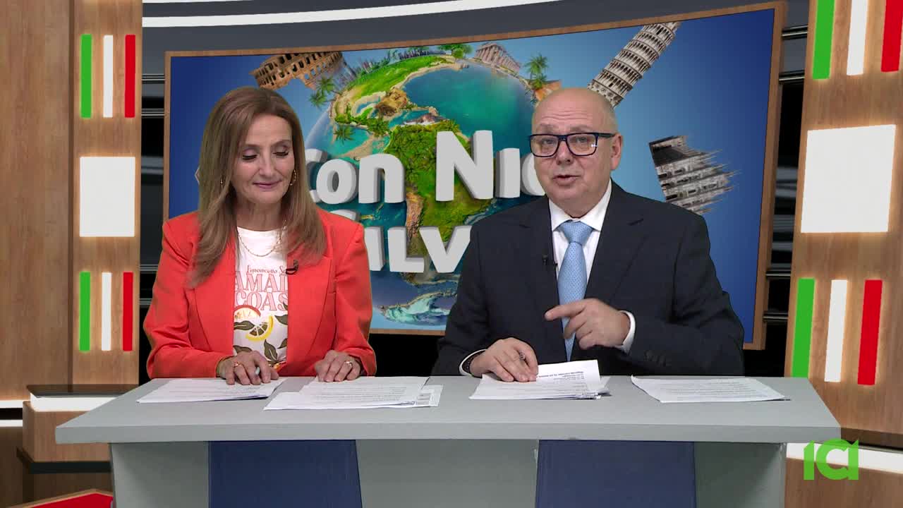 The man in the suit gestures with his right hand, speaking directly to the camera. Beside him, the woman in the orange blazer smiles while looking down at her papers.
