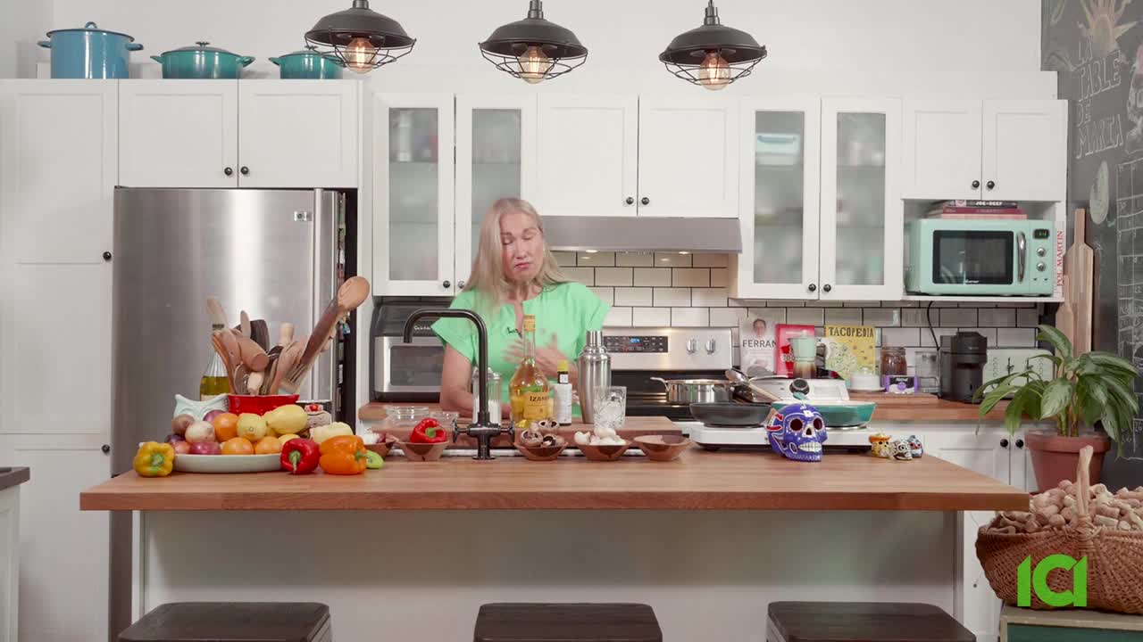 A woman in a green shirt gestures as she speaks, framed by a kitchen counter laden with ingredients and cooking implements. The scene is lit by overhead lights, likely for CFHD-DT in Canada.
