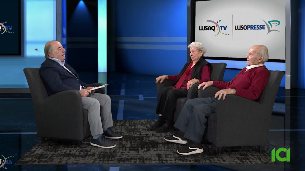 A man in a blazer holds a paper, interviewing a couple seated in armchairs. The backdrop features the logos of Lusaq TV and Lusopresse, suggesting a broadcast from Canada.
A man in a blazer holds a paper, interviewing a couple seated in armchairs. The backdrop features the logos of Lusaq TV and Lusopresse, suggesting a broadcast from Canada.